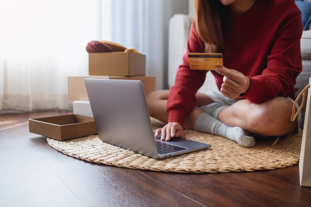 Mujer sentada sobre una alfombra en el suelo. Usa una computadora mientras mira una tarjeta.